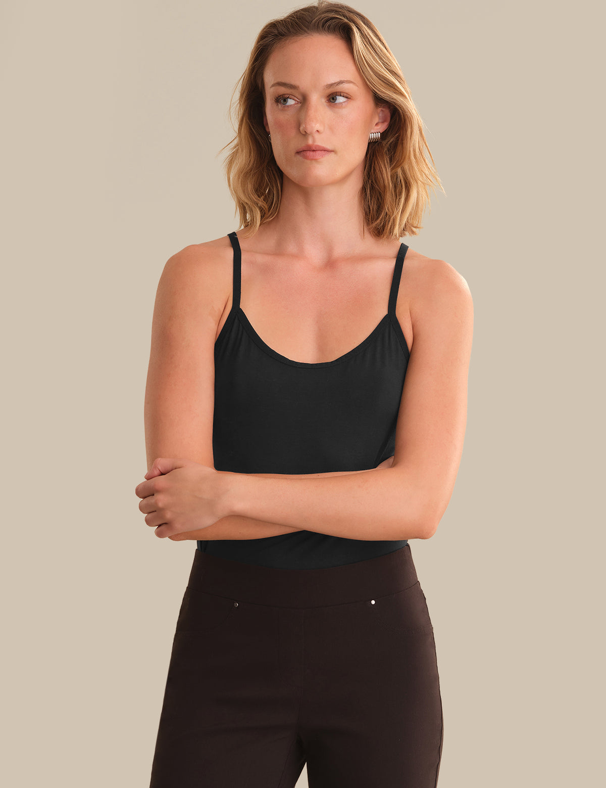 A woman modeling a black spaghetti-strap camisole tank top paired with dark brown pants, set against a solid beige studio background.