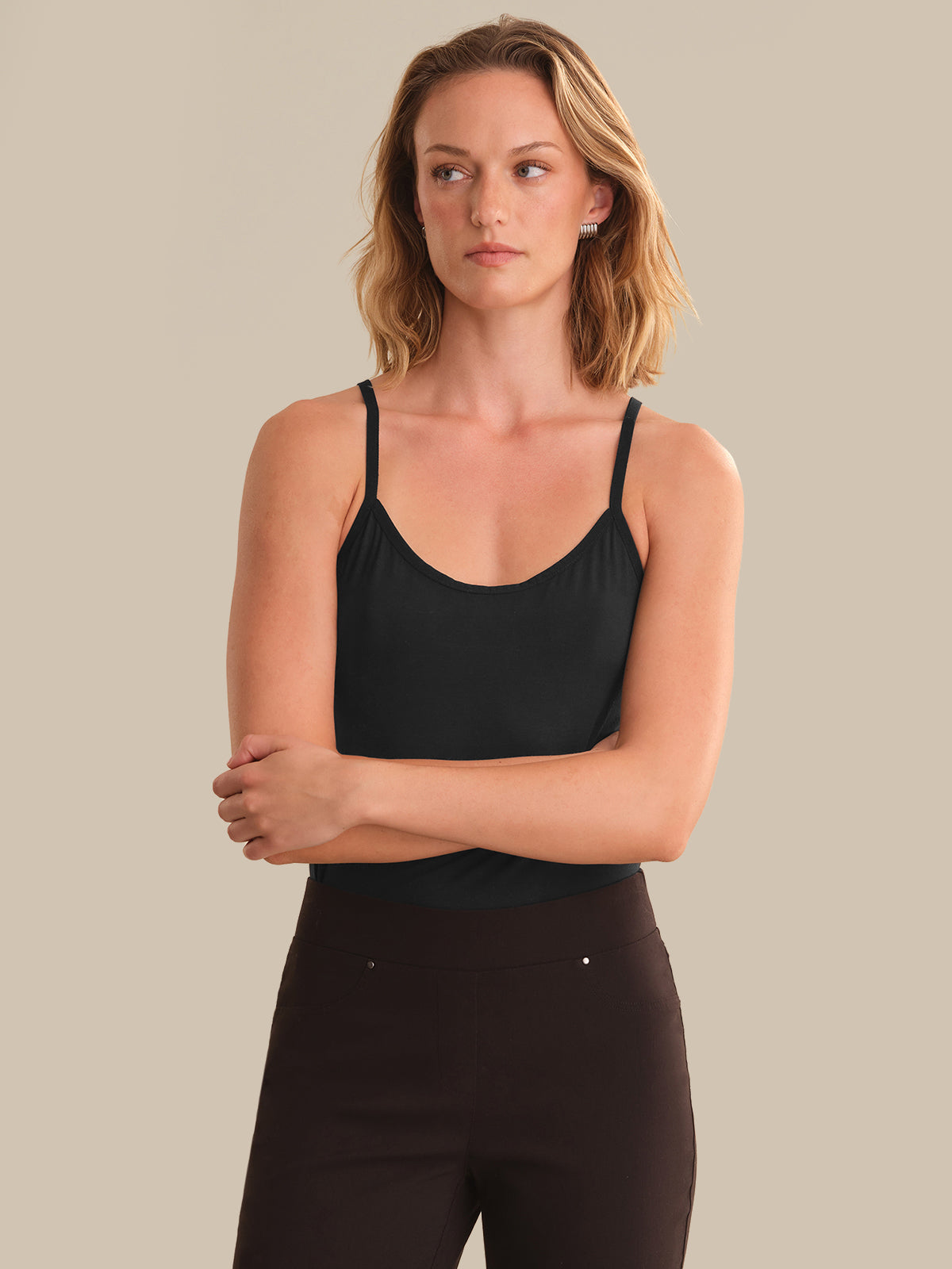A woman modeling a black spaghetti-strap camisole tank top paired with dark brown pants, set against a solid beige studio background.