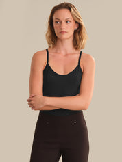 A woman modeling a black spaghetti-strap camisole tank top paired with dark brown pants, set against a solid beige studio background.