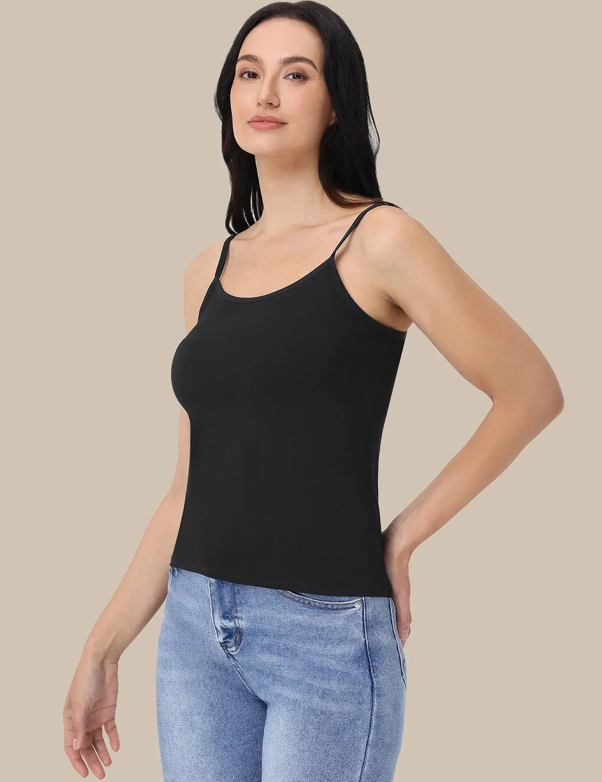 A woman modeling a black spaghetti-strap tank top paired with light blue jeans, set against a solid beige background.