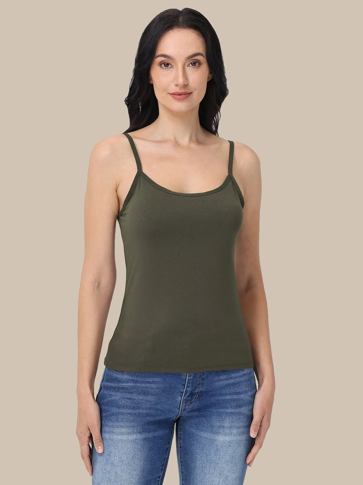 A woman modeling an olive green spaghetti-strap tank top paired with blue jeans, set against a solid beige background.