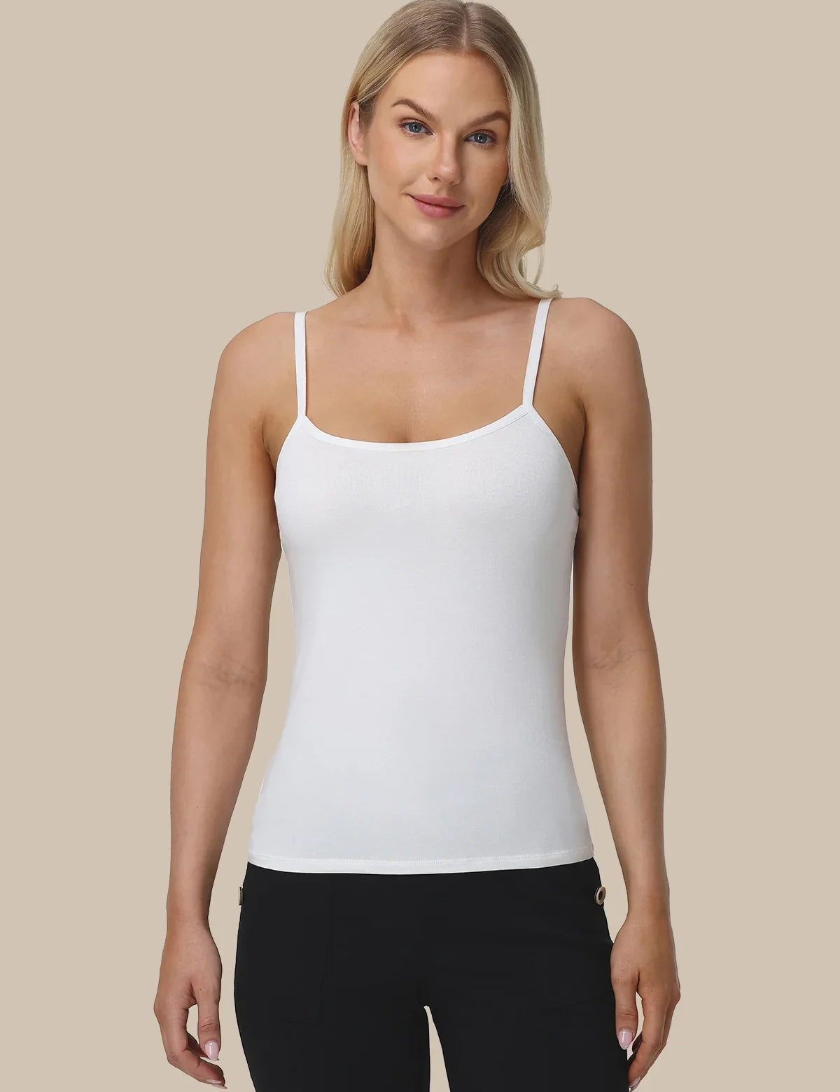 A woman modeling a white spaghetti-strap tank top with black pants, standing against a solid beige background.