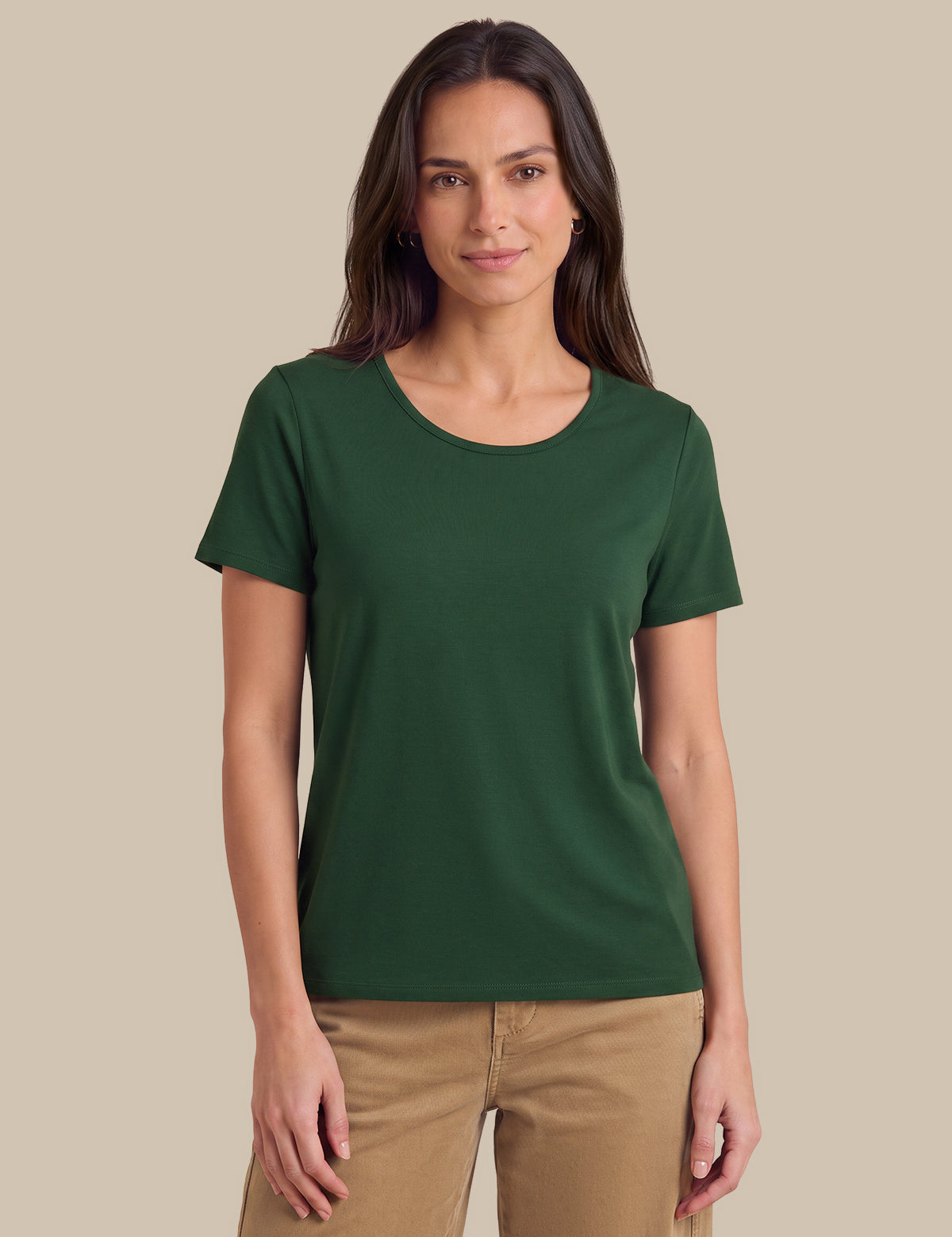 A woman modeling a dark green short-sleeve crewneck t-shirt with tan pants, standing against a solid beige studio background.