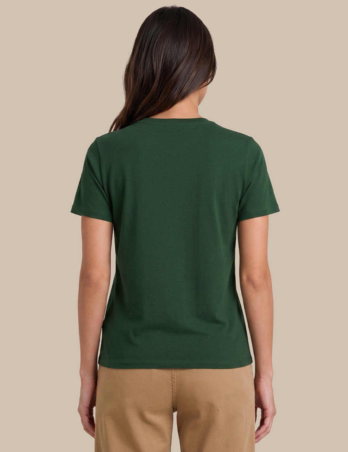 A woman viewed from the back wearing a dark green short-sleeve crewneck t-shirt and tan pants, set against a solid beige background.