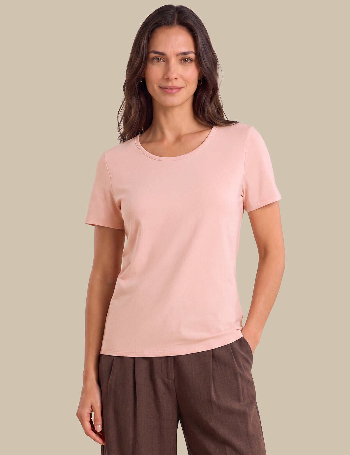 A woman modeling a light pink short-sleeve crewneck t-shirt with dark brown trousers, standing against a solid beige background.