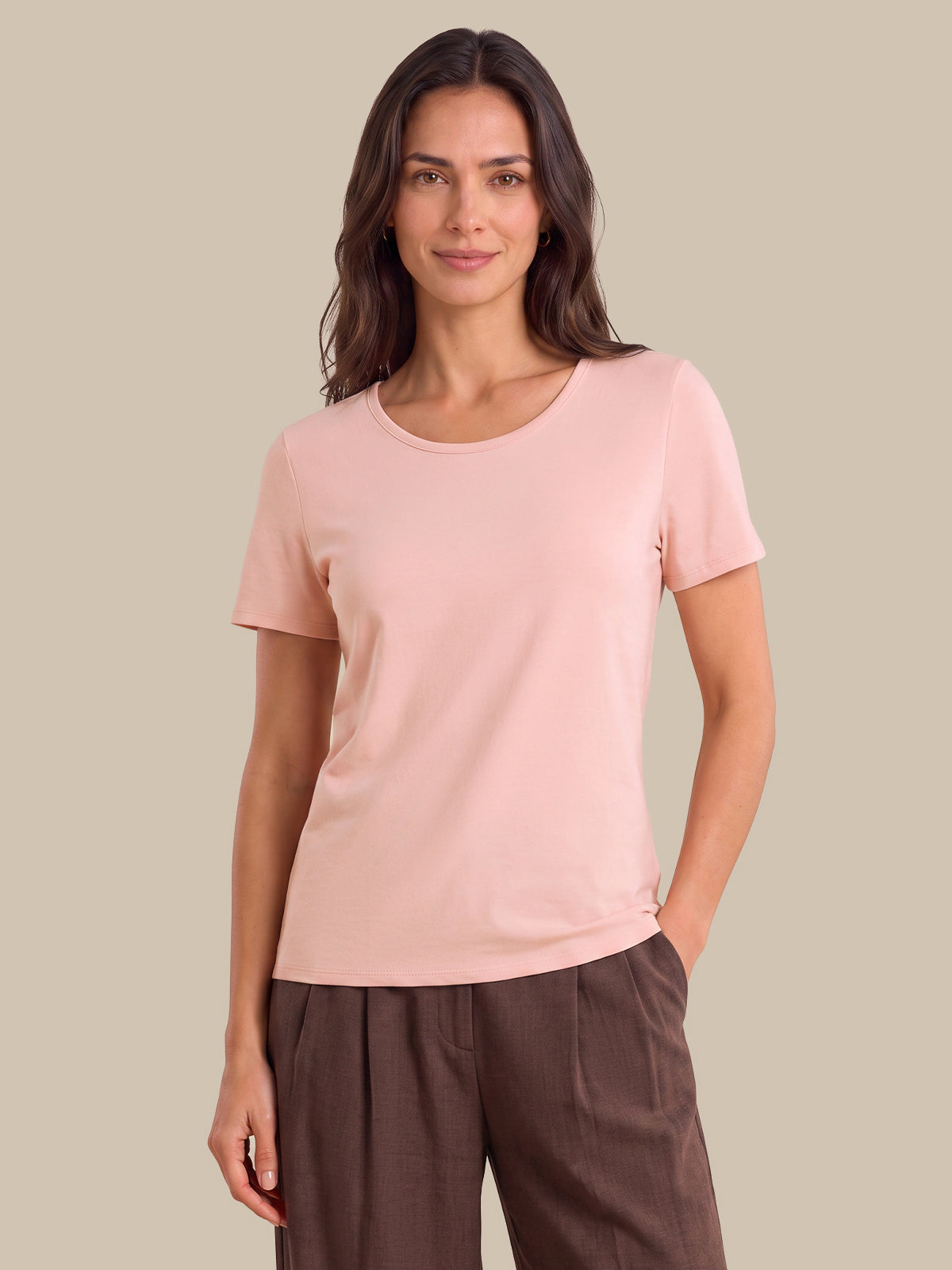 A woman modeling a light pink short-sleeve crewneck t-shirt with dark brown trousers, standing against a solid beige background.