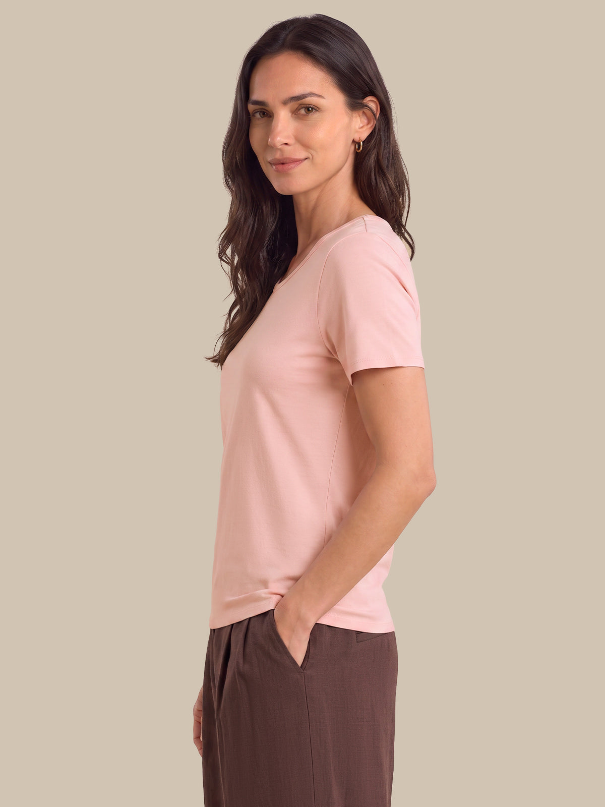 A woman modeling a light pink short-sleeve V-neck top, paired with brown trousers, against a solid beige background.