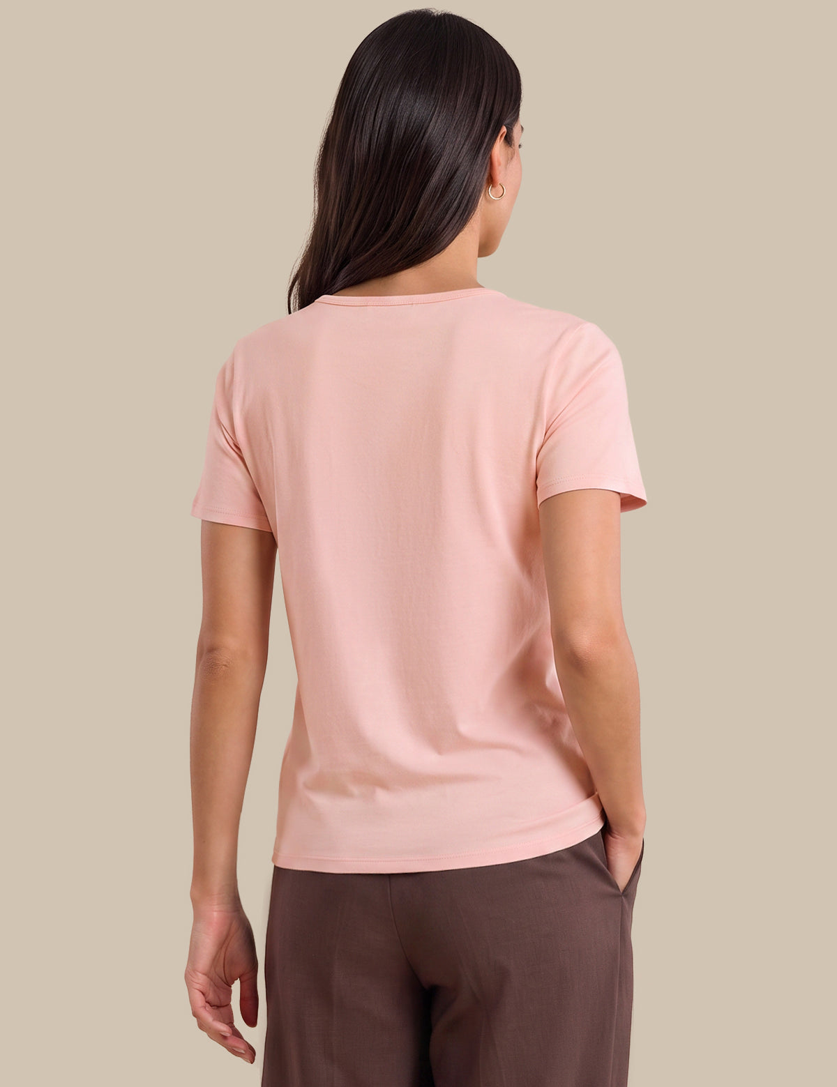 A woman shown from the back wearing a light pink short-sleeve crewneck t-shirt and dark brown trousers, set against a solid beige studio background.