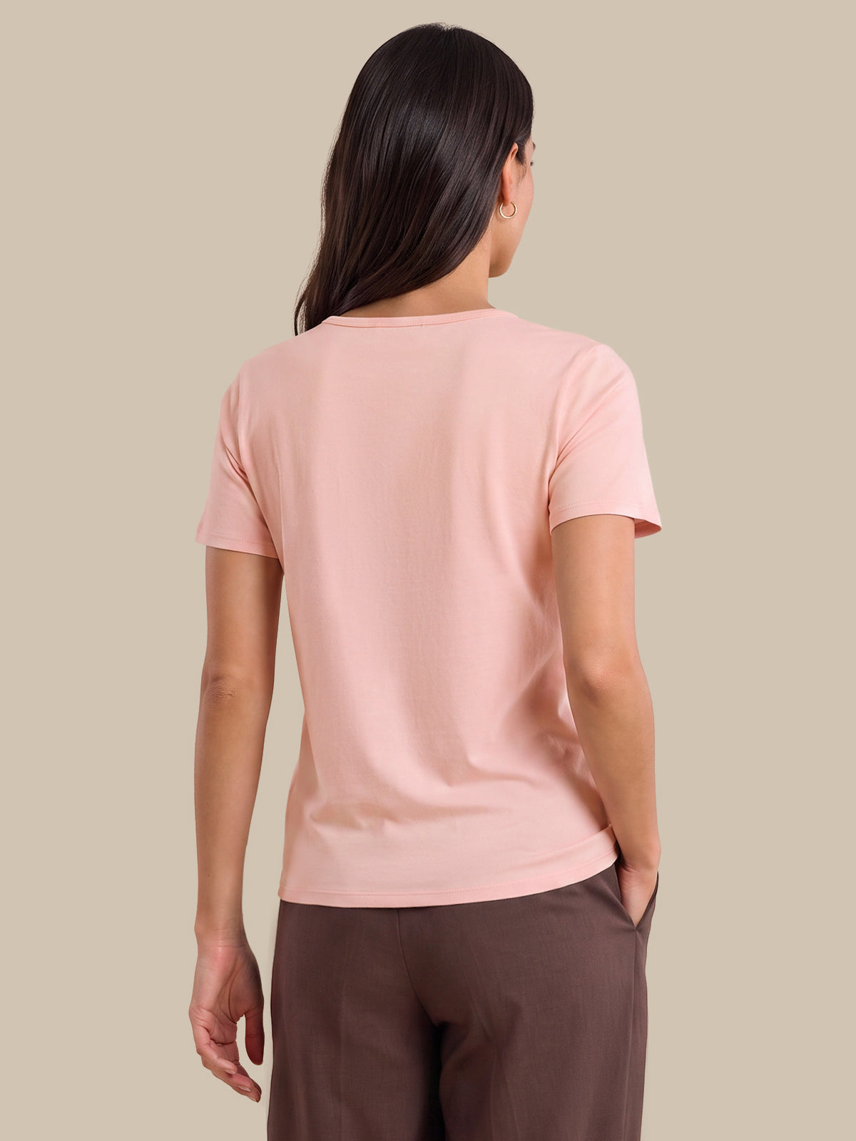 A woman shown from the back wearing a light pink short-sleeve crewneck t-shirt and dark brown trousers, set against a solid beige studio background.