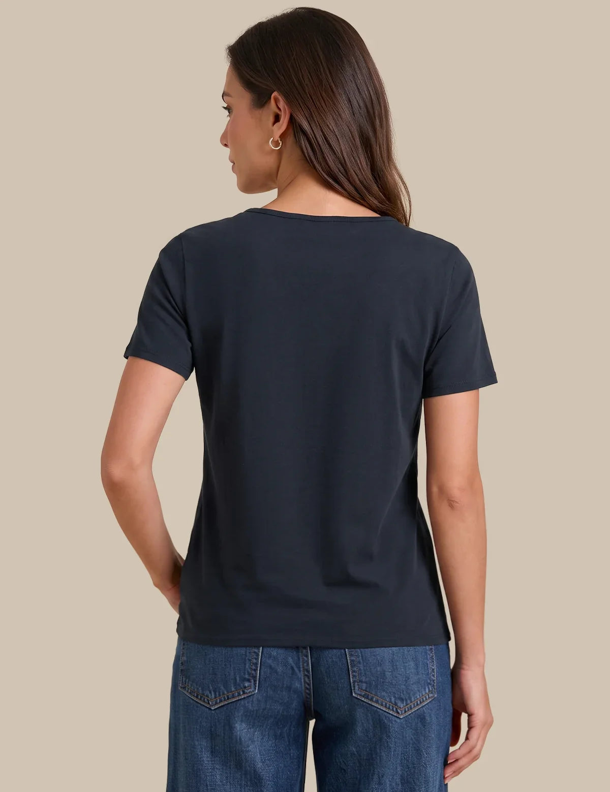 A woman shown from the back wearing a dark navy blue short-sleeve crewneck t-shirt and blue jeans, set against a solid beige background.