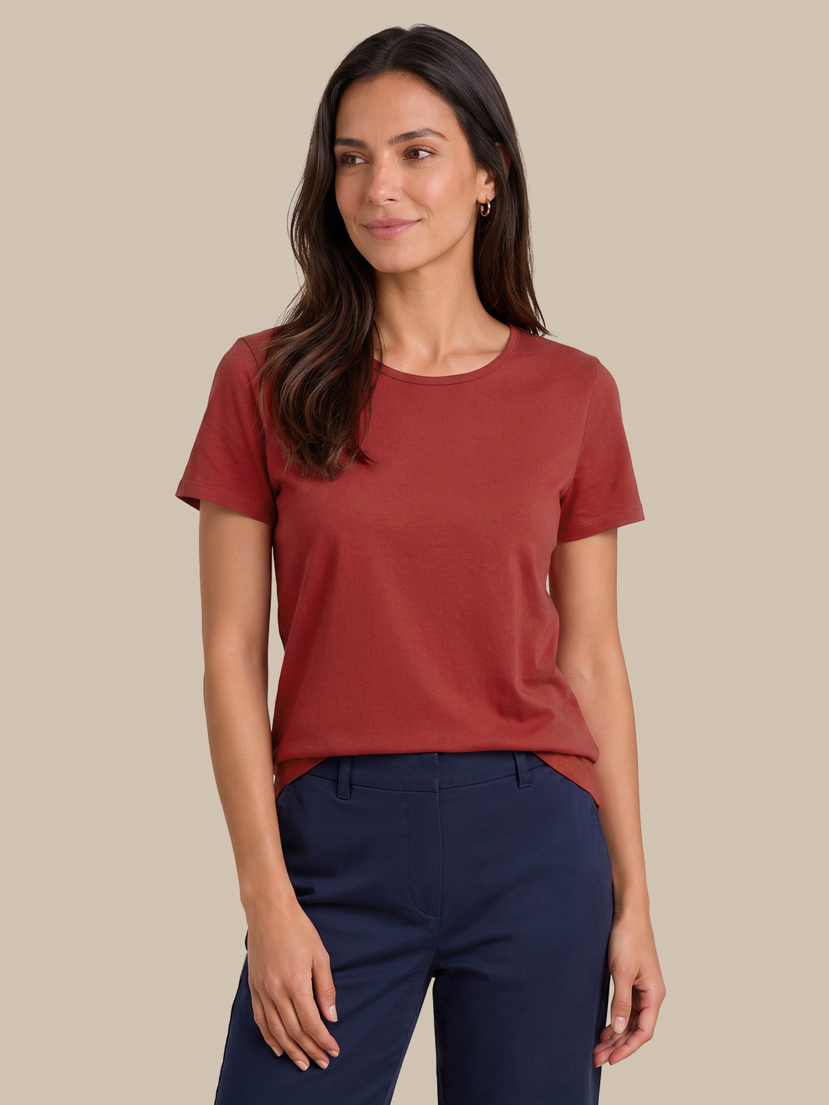A woman modeling a short-sleeve crewneck t-shirt in a muted terracotta or rust color, paired with navy blue pants, against a solid beige background.