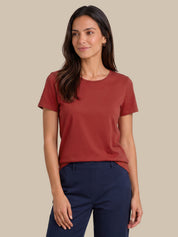 A woman modeling a short-sleeve crewneck t-shirt in a muted terracotta or rust color, paired with navy blue pants, against a solid beige background.