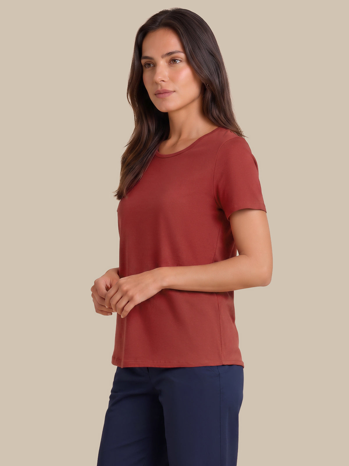 A woman modeling a short-sleeve crewneck top in a muted terracotta or rust color, paired with navy pants, against a solid beige background.