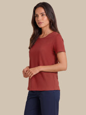 A woman modeling a short-sleeve crewneck top in a muted terracotta or rust color, paired with navy pants, against a solid beige background.