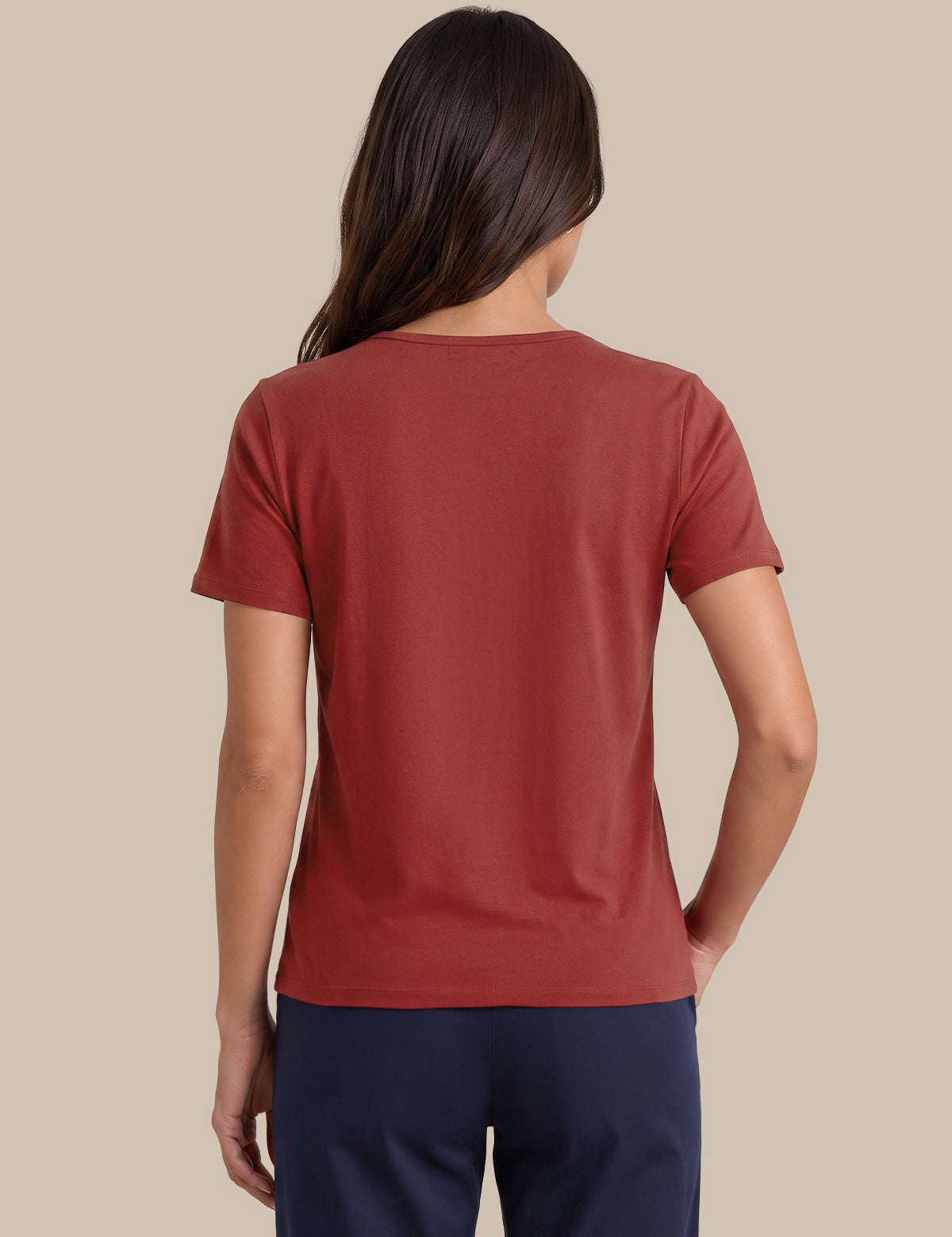 A woman viewed from the back wearing a rust-colored short-sleeve crewneck t-shirt and navy blue pants, set against a solid beige background.
