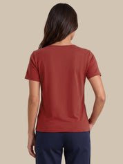 A woman viewed from the back wearing a rust-colored short-sleeve crewneck t-shirt and navy blue pants, set against a solid beige background.