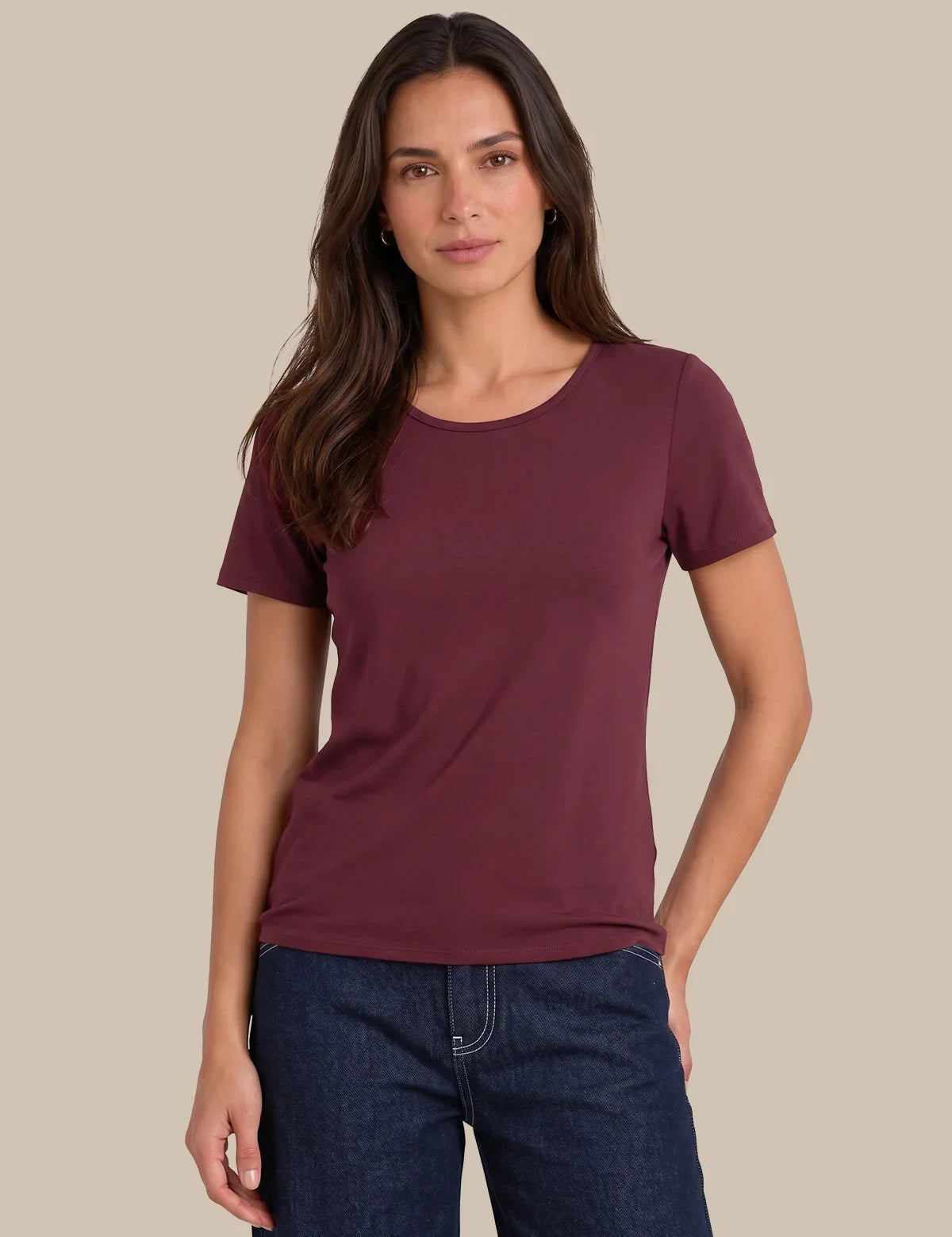 A woman modeling a short-sleeve crewneck t-shirt in a deep burgundy color, paired with dark blue jeans, against a solid beige background.