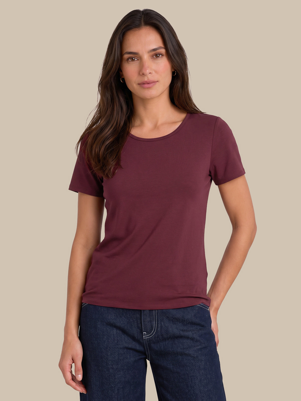 A woman modeling a short-sleeve crewneck t-shirt in a deep burgundy color, paired with dark blue jeans, against a solid beige background.