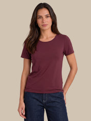 A woman modeling a short-sleeve crewneck t-shirt in a deep burgundy color, paired with dark blue jeans, against a solid beige background.