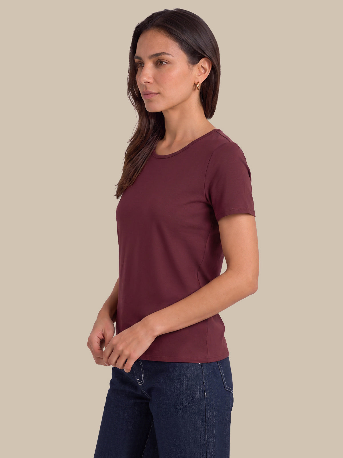 A woman wearing a short-sleeve crew-neck t-shirt in maroon and dark blue jeans, standing against a solid beige background.