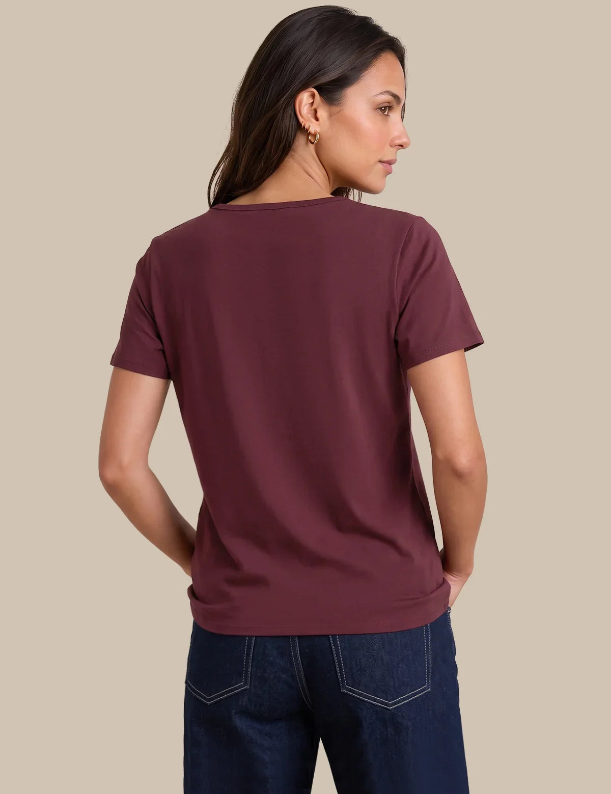 A woman shown from the back wearing a short-sleeve crew-neck t-shirt in a deep burgundy color, paired with dark blue jeans, against a solid beige background.