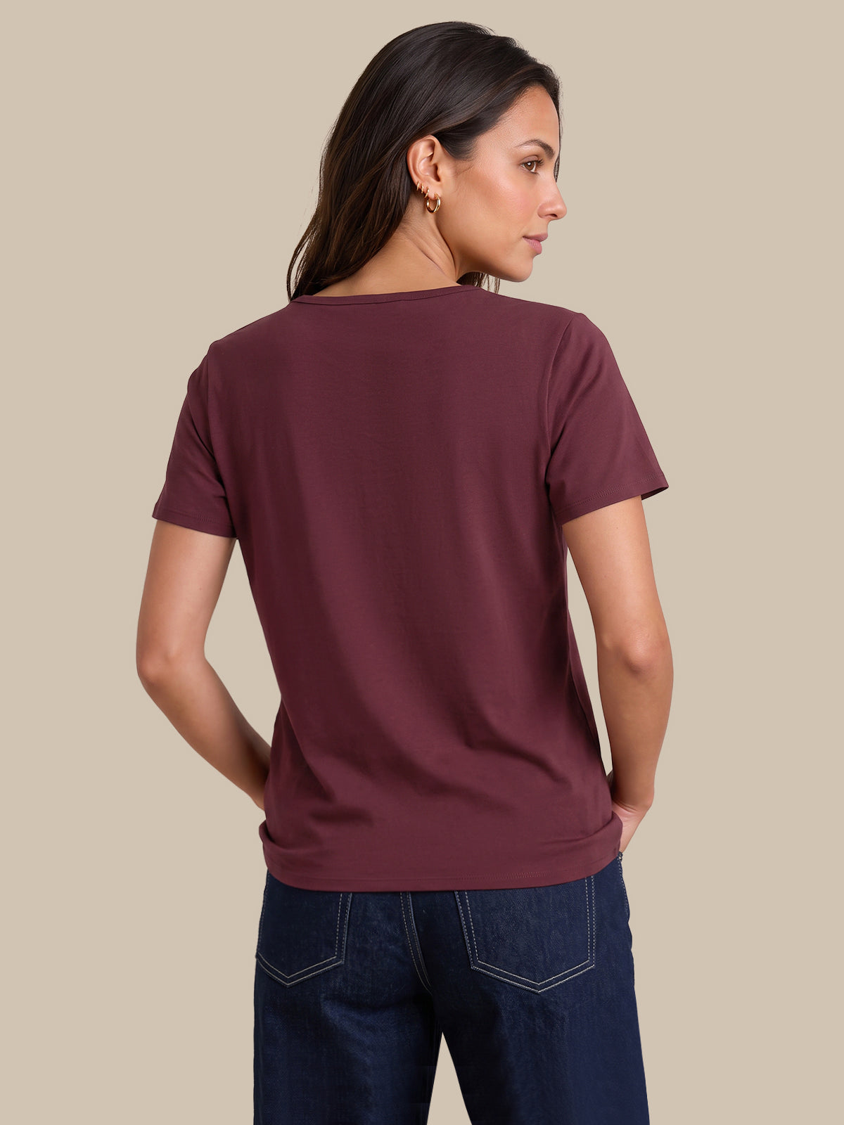 A woman shown from the back wearing a short-sleeve crew-neck t-shirt in a deep burgundy color, paired with dark blue jeans, against a solid beige background.