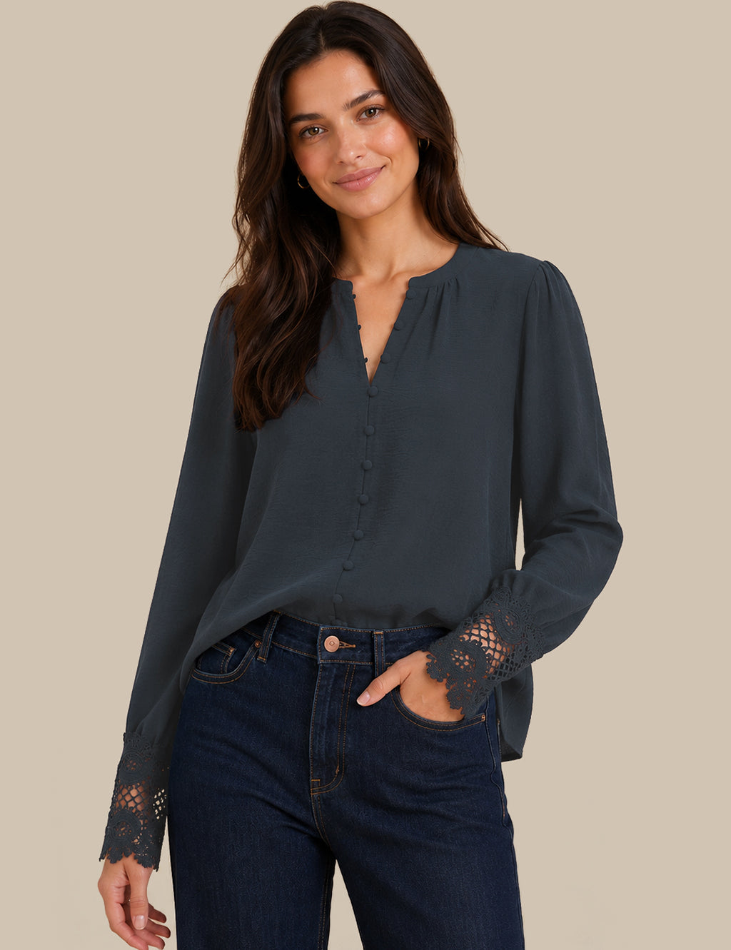 A woman modeling a dark slate blue long-sleeve blouse with lace detailing on the cuffs and a split neckline, paired with dark blue jeans, against a solid beige background.