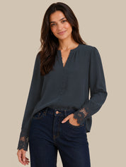 A woman modeling a dark slate blue long-sleeve blouse with lace detailing on the cuffs and a split neckline, paired with dark blue jeans, against a solid beige background.