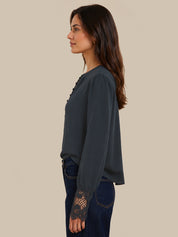 A woman shown in side profile wearing a dark slate blue blouse with lace detailing on the cuffs and chest, paired with dark blue jeans, against a solid beige background.