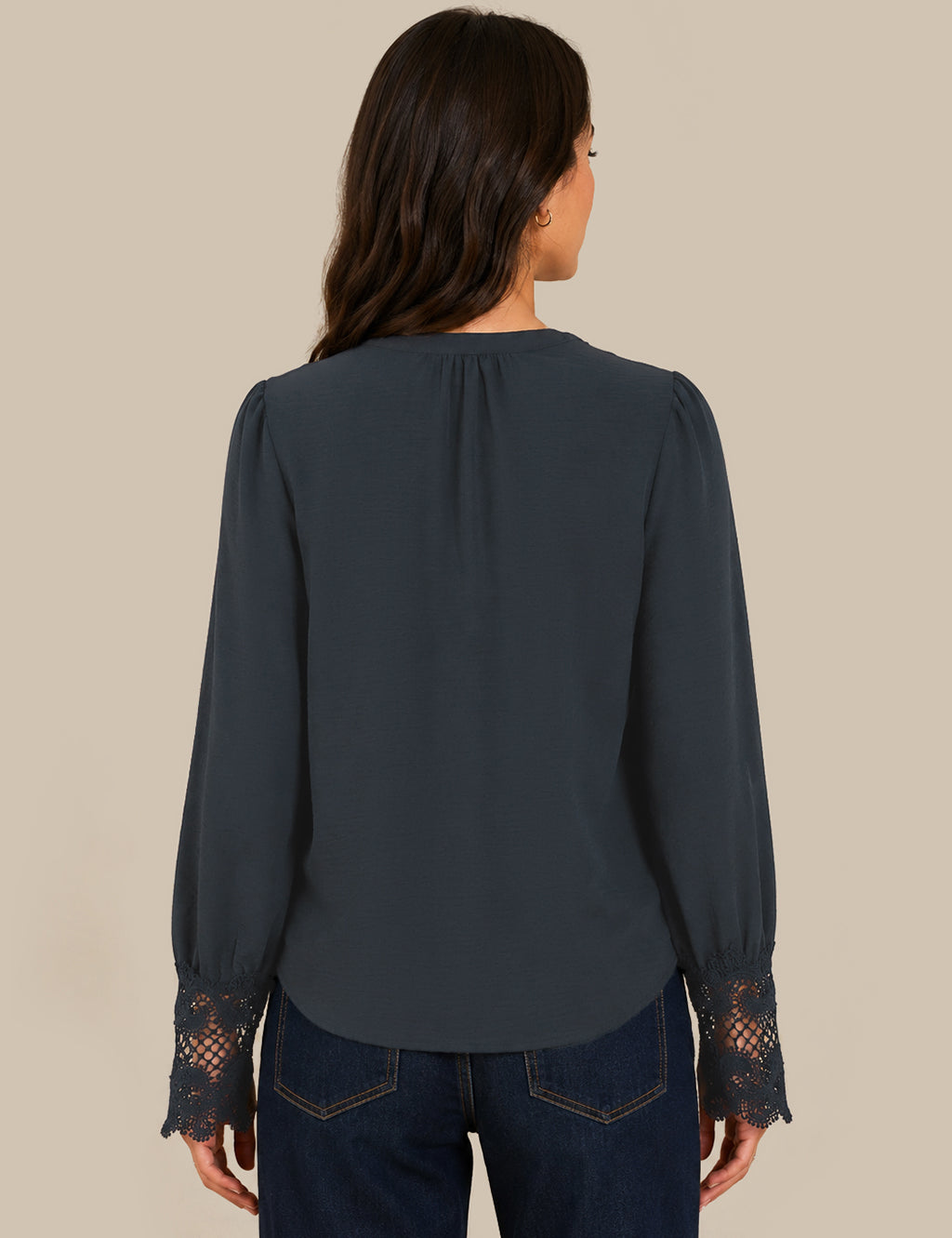 A woman viewed from the back wearing a dark slate blue blouse with lace cuffs and dark blue jeans, set against a solid beige background.