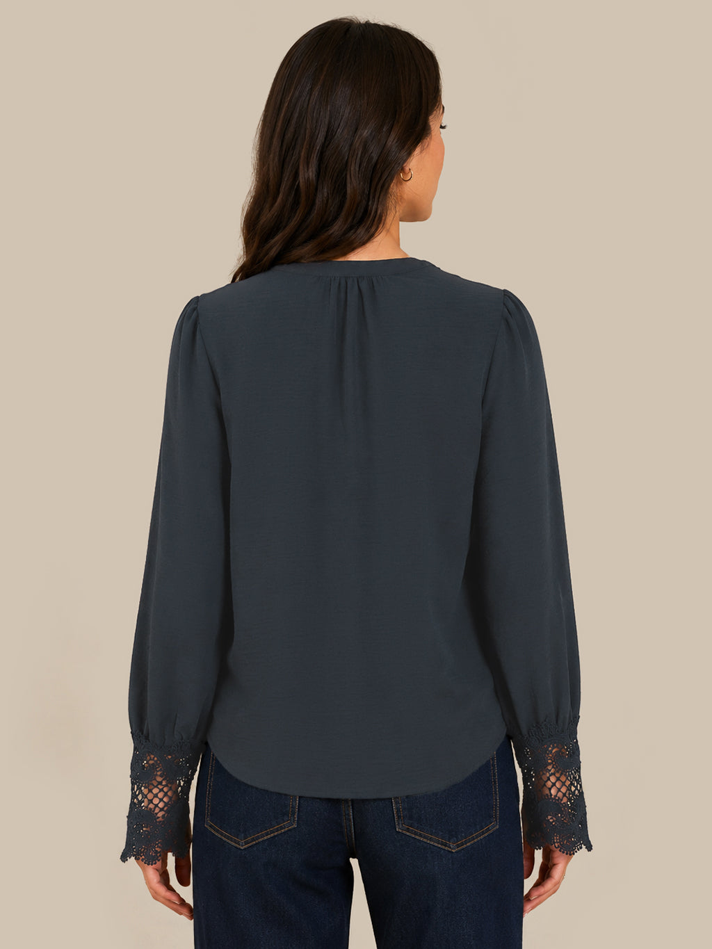A woman viewed from the back wearing a dark slate blue blouse with lace cuffs and dark blue jeans, set against a solid beige background.