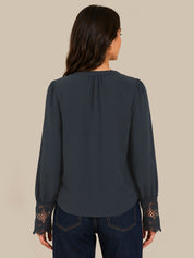 A woman viewed from the back wearing a dark slate blue blouse with lace cuffs and dark blue jeans, set against a solid beige background.