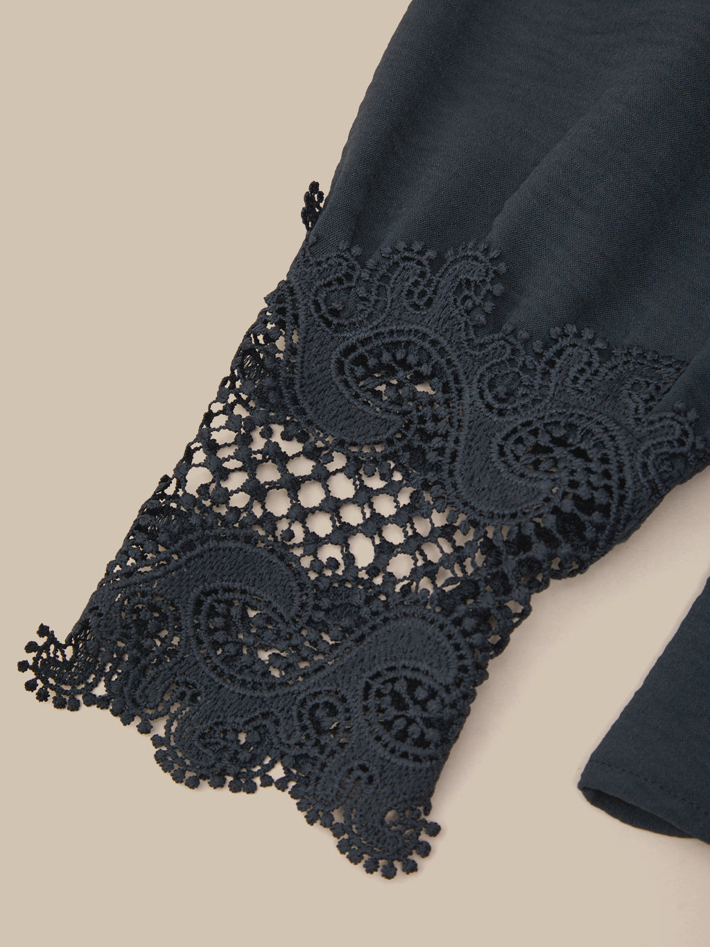 Close-up of a dark navy blue garment featuring an intricate, matching lace cuff with a scalloped and floral pattern, set against a solid beige background.