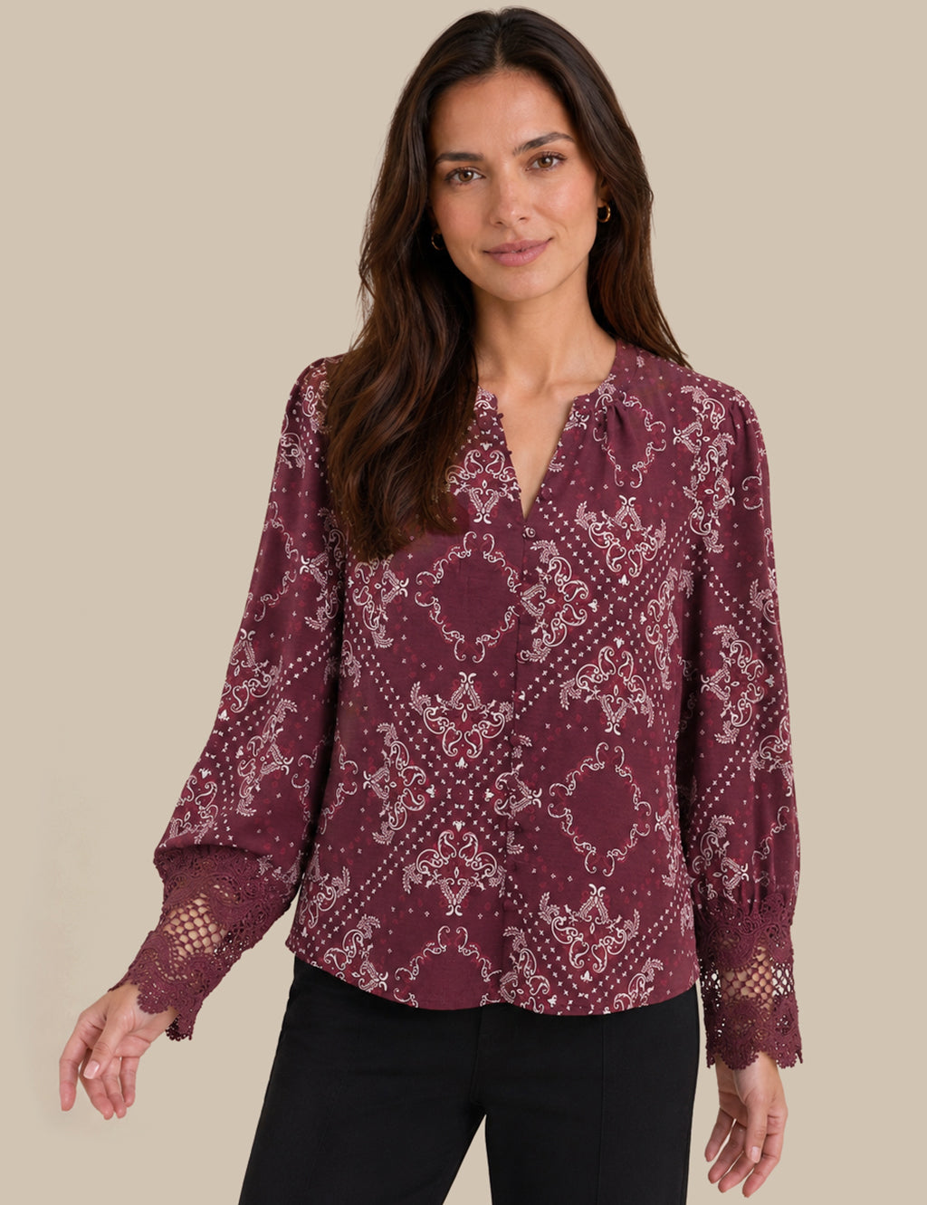 A woman wearing a long-sleeve burgundy blouse with a white damask pattern and lace cuffs, paired with black pants, against a solid beige background.