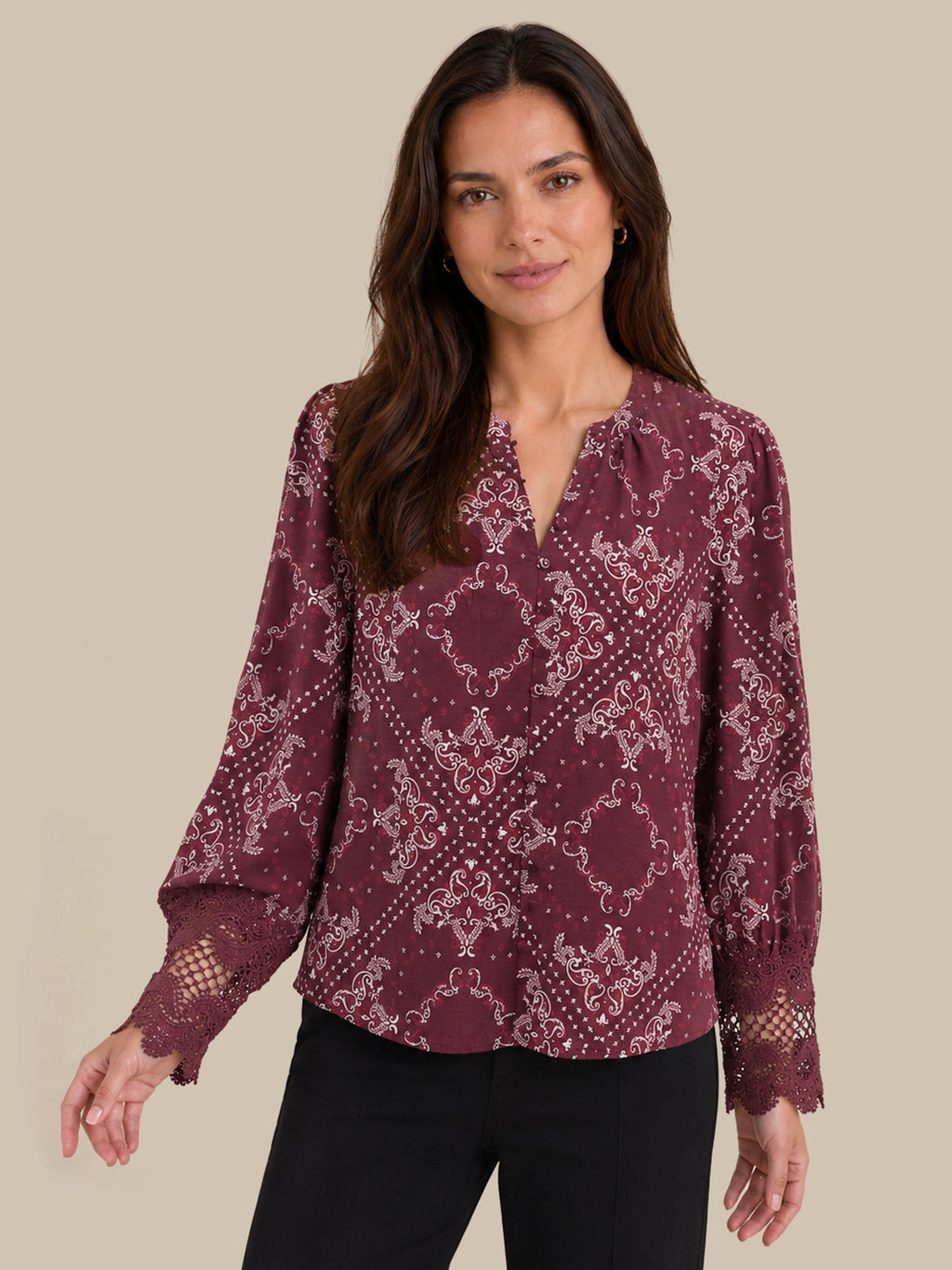 A woman wearing a long-sleeve burgundy blouse with a white damask pattern and lace cuffs, paired with black pants, against a solid beige background.