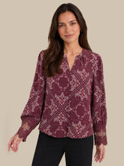 A woman wearing a long-sleeve burgundy blouse with a white damask pattern and lace cuffs, paired with black pants, against a solid beige background.