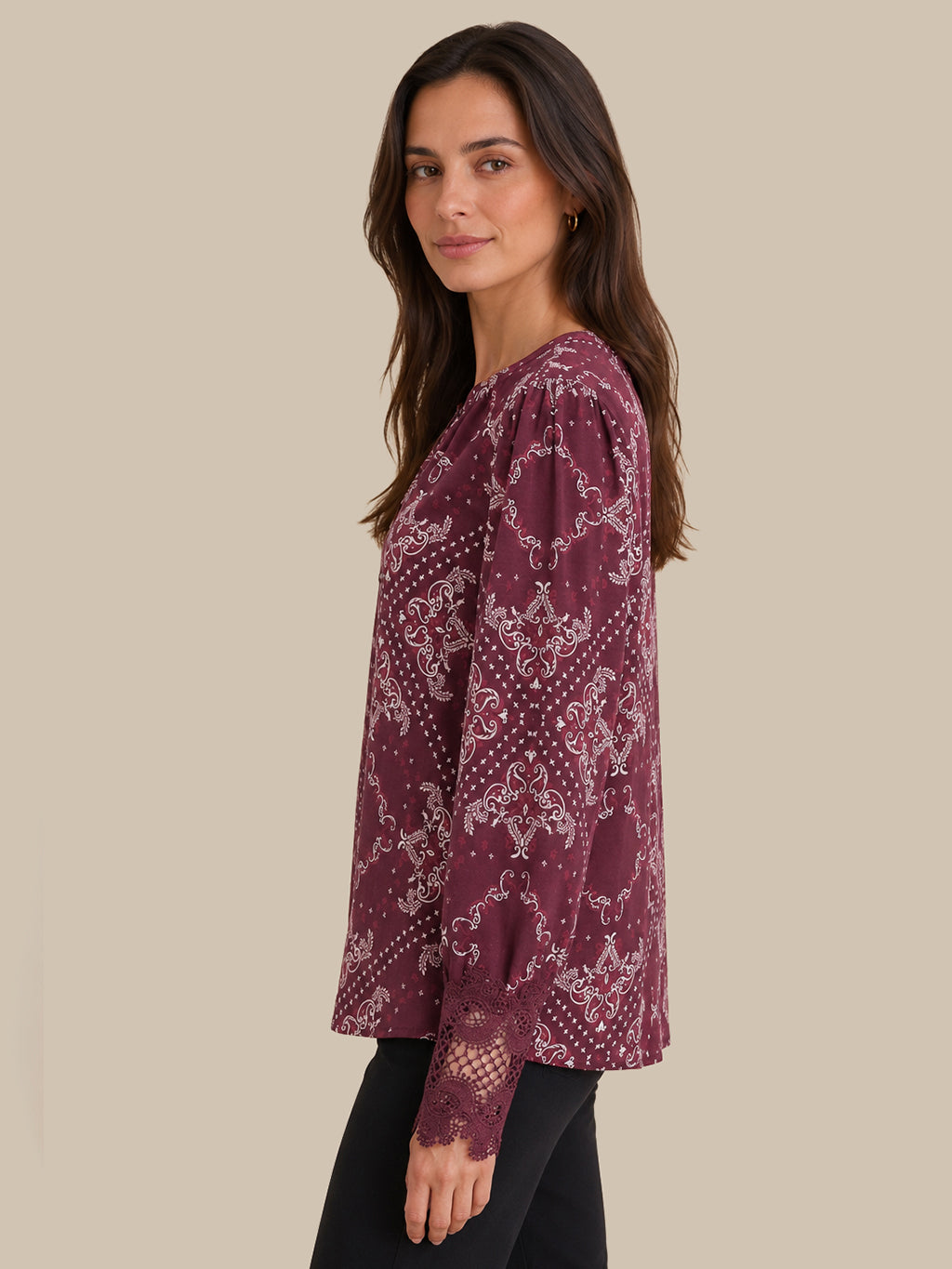 A woman wearing a long-sleeve blouse with a dusty rose and white paisley pattern, paired with black pants, shown from a side-back angle against a solid beige background.