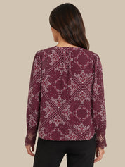 A woman shown from the back wearing a long-sleeve blouse with a burgundy and white paisley pattern, paired with black pants, against a solid beige background.