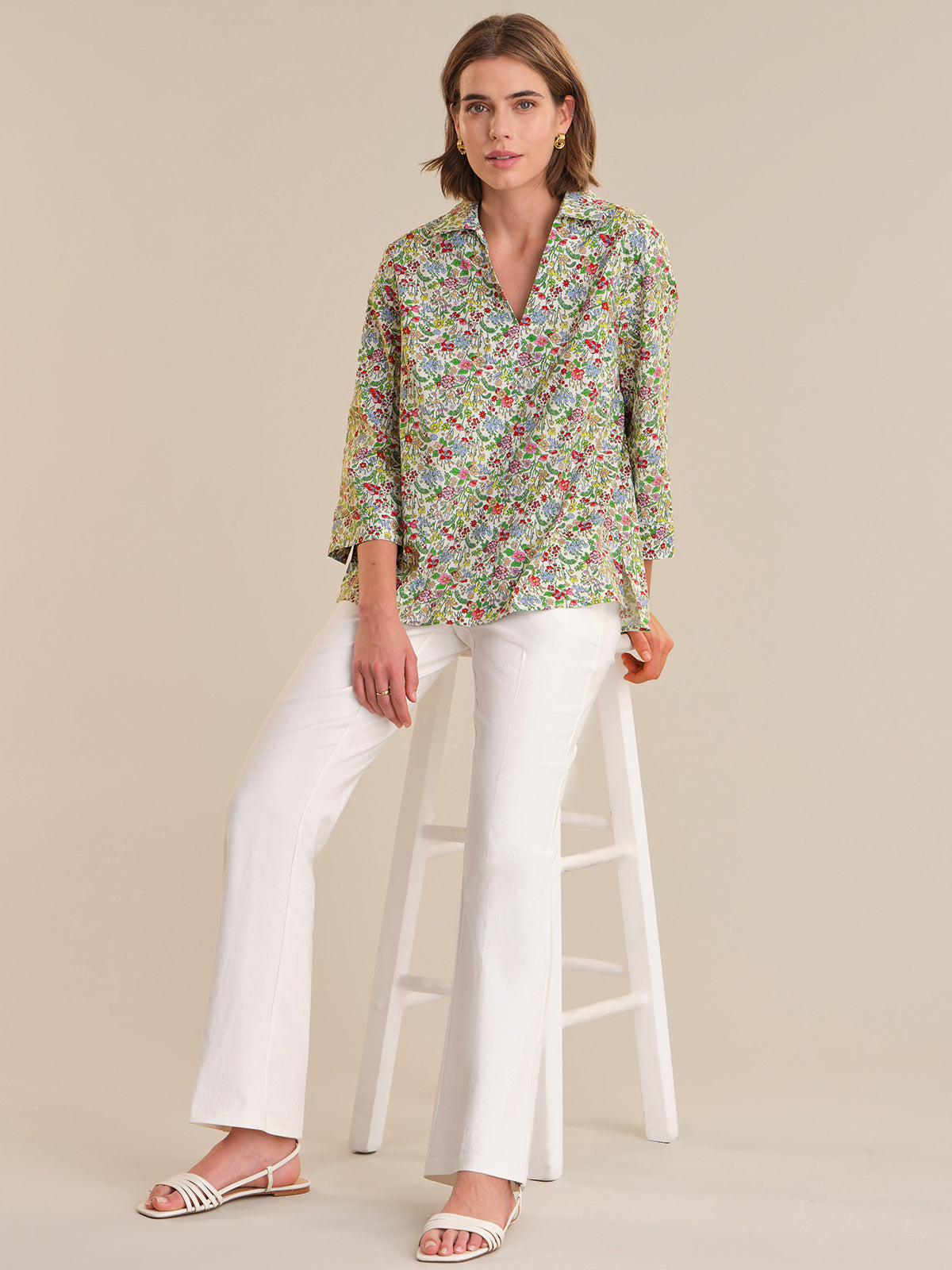 Woman sitting on a stool wearing a floral blouse and white pants against a beige background