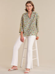Woman sitting on a stool wearing a floral blouse and white pants against a beige background