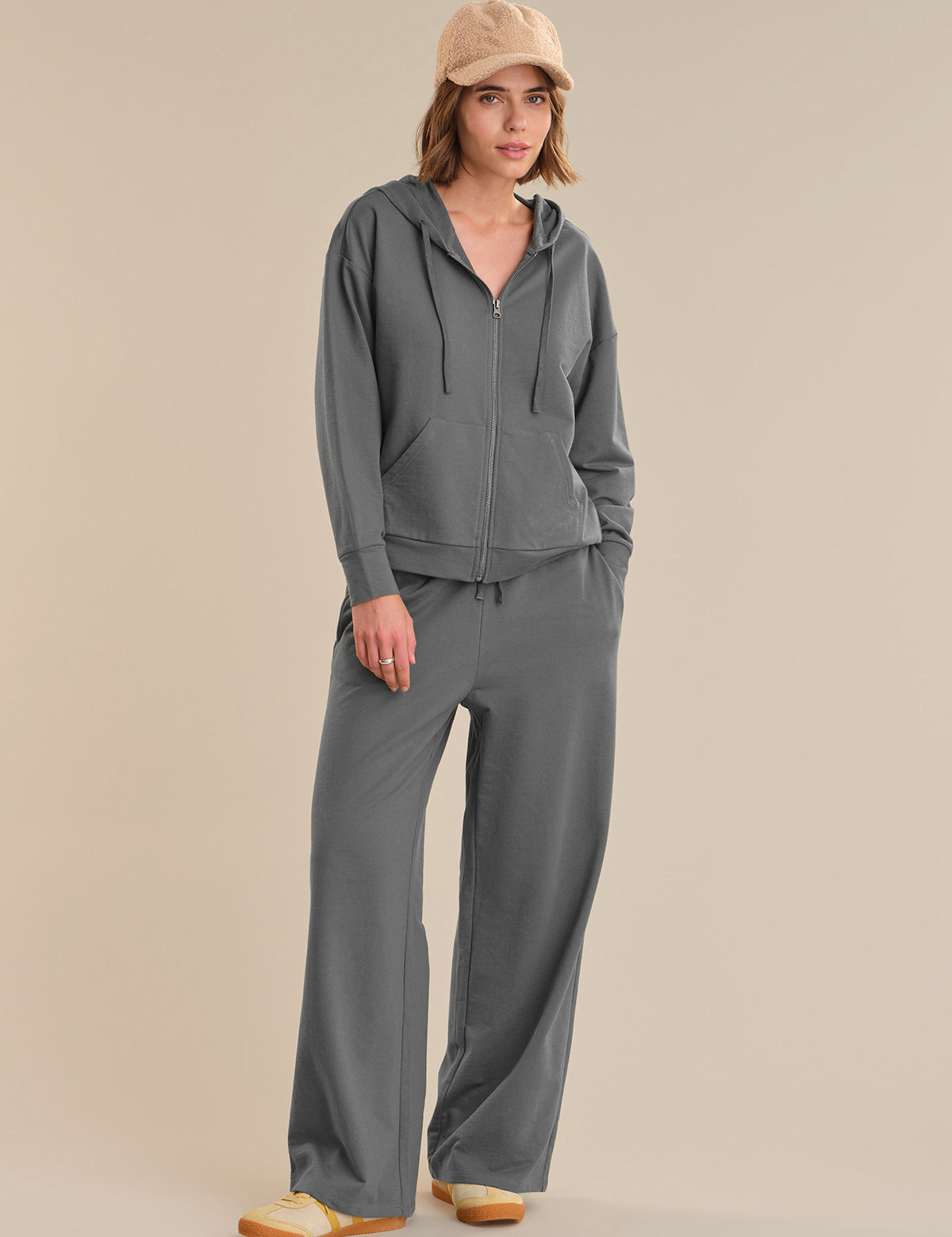 Woman wearing a gray hoodie and pants with a beige cap on a beige background