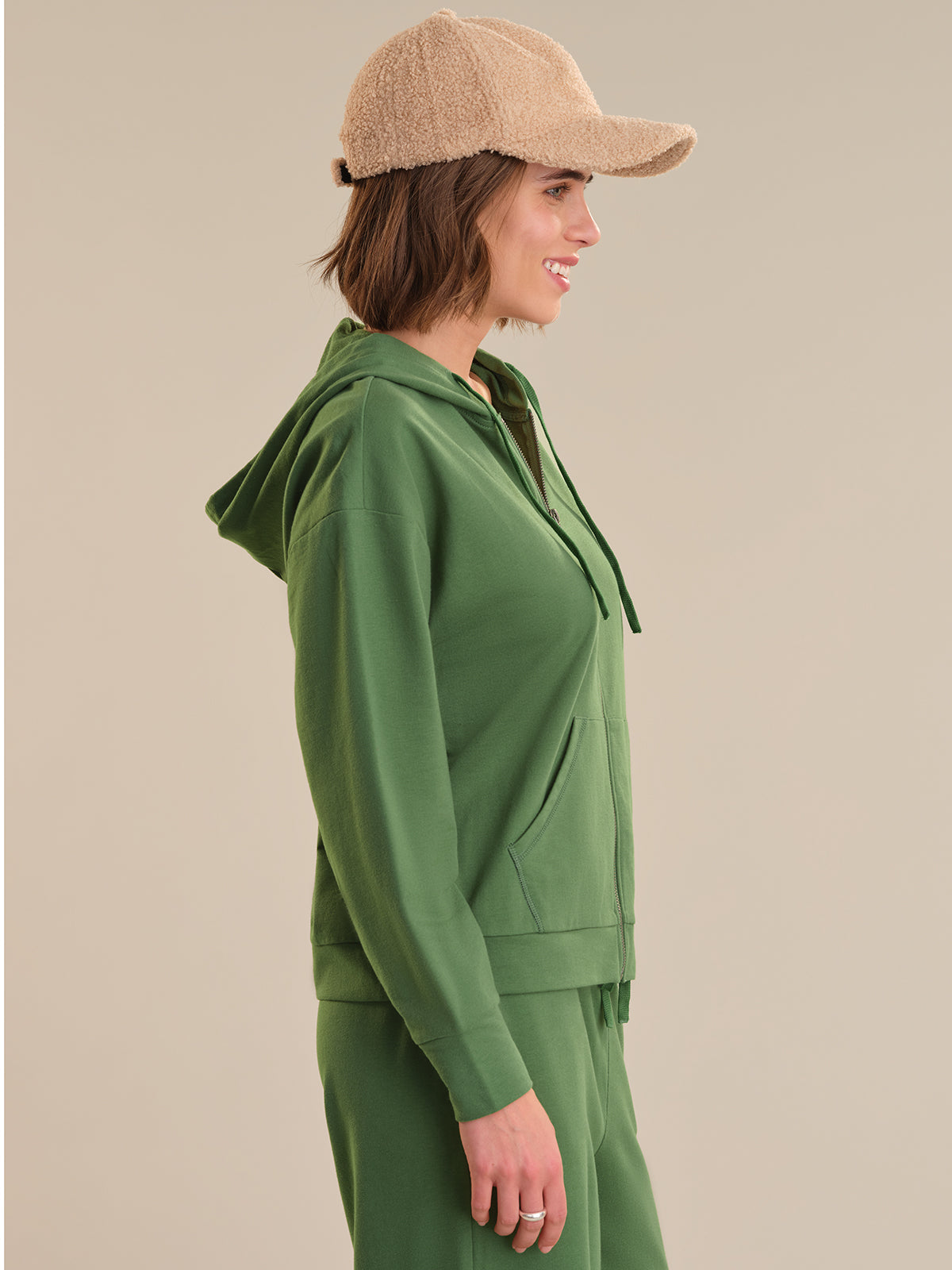 Woman wearing a green hoodie and matching pants with a beige cap on a beige background