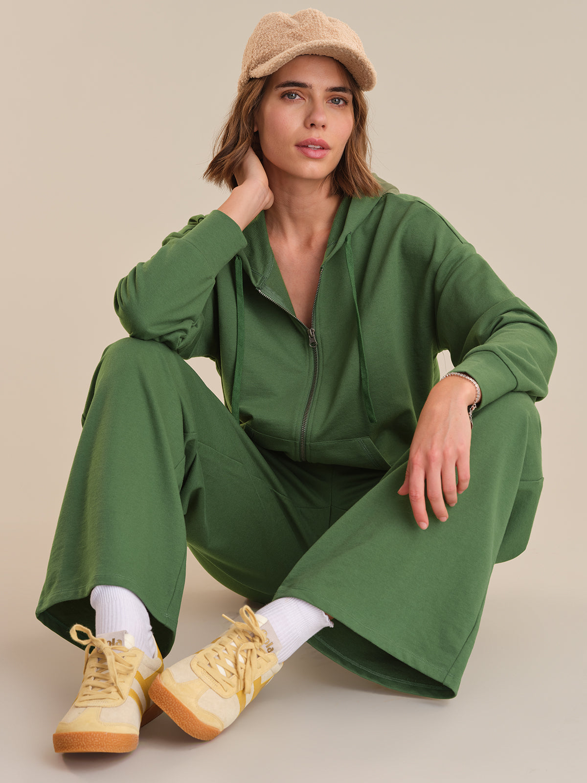 Woman wearing a green tracksuit with beige shoes and cap on a neutral background