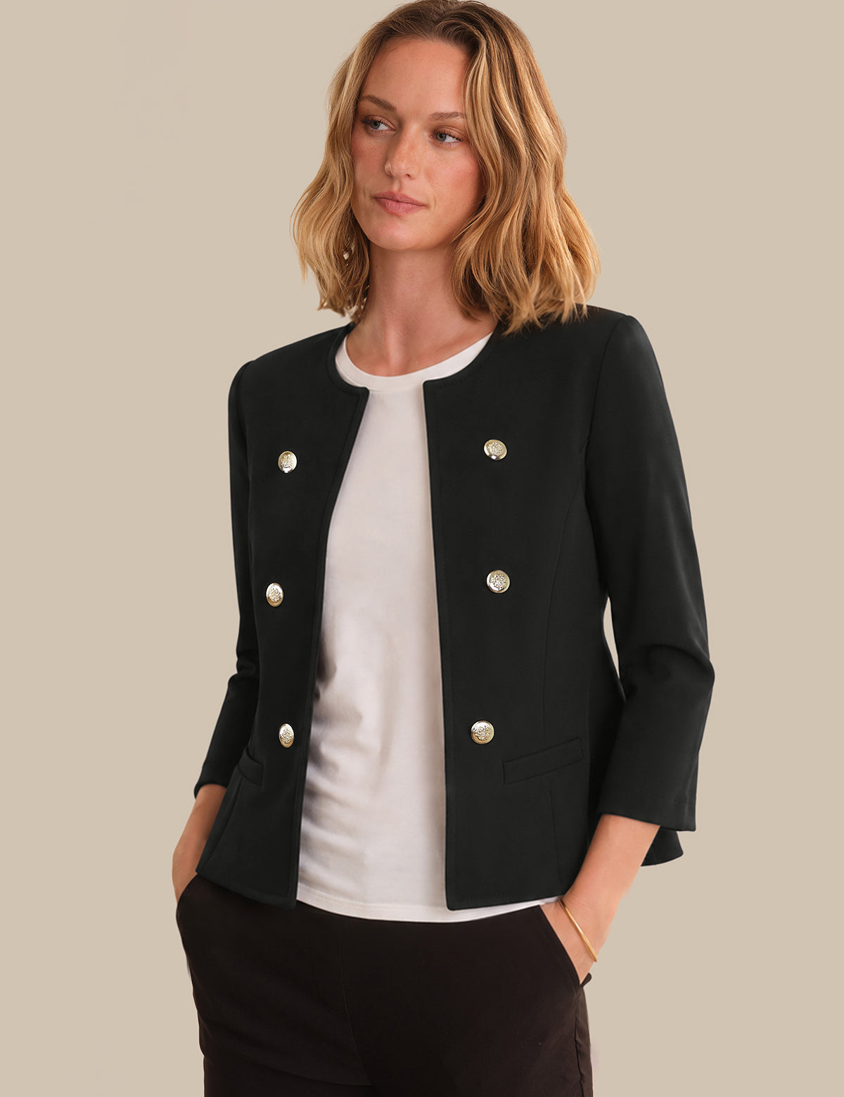 A woman wearing a black double-breasted blazer with gold buttons over a white top and black trousers, photographed against a solid beige background.