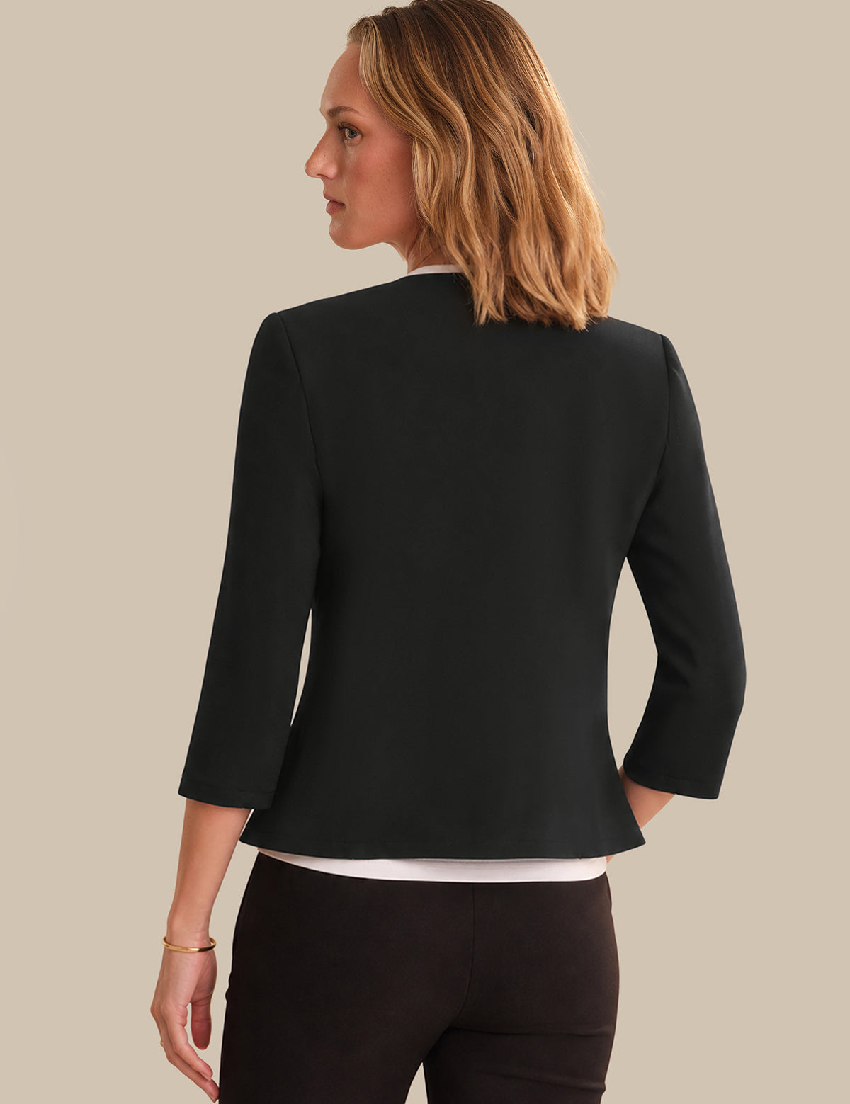 A woman shown from the back wearing a black three-quarter sleeve blazer over a white top and dark pants, against a solid beige background.