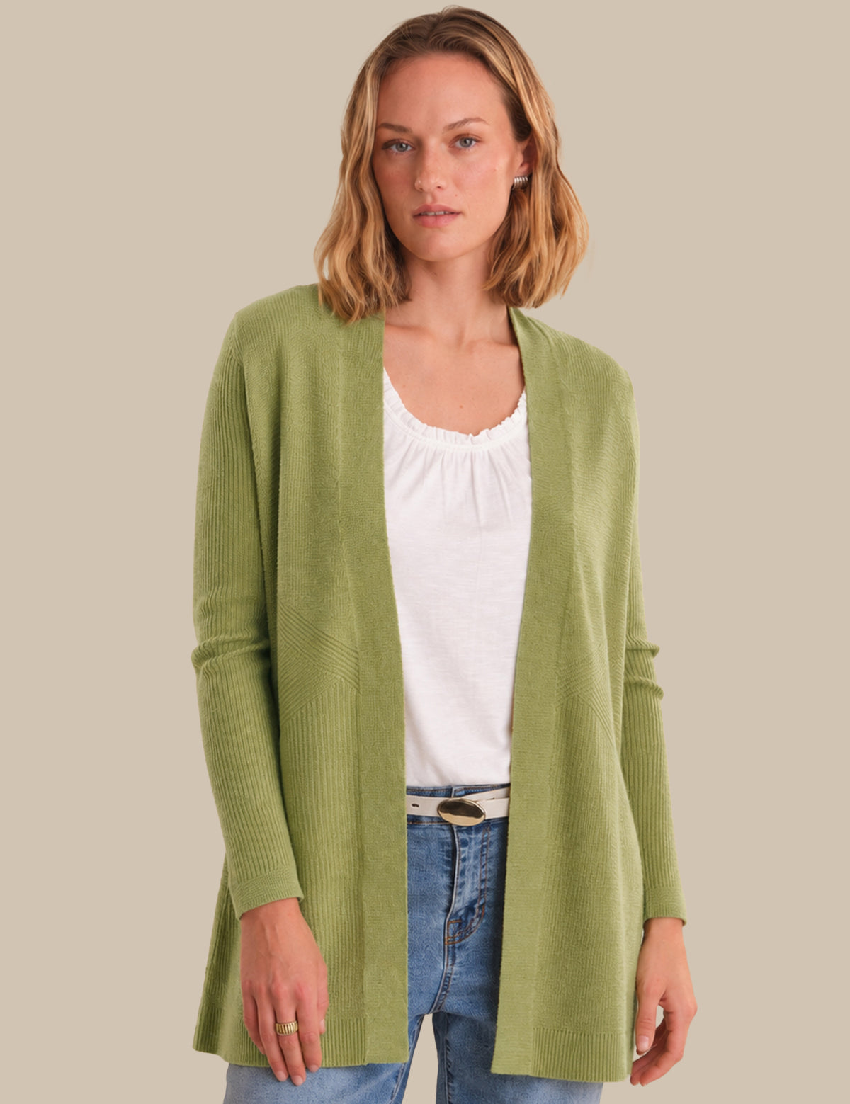 A woman modeling a light olive green open-front cardigan over a white top and blue jeans, set against a solid beige background.