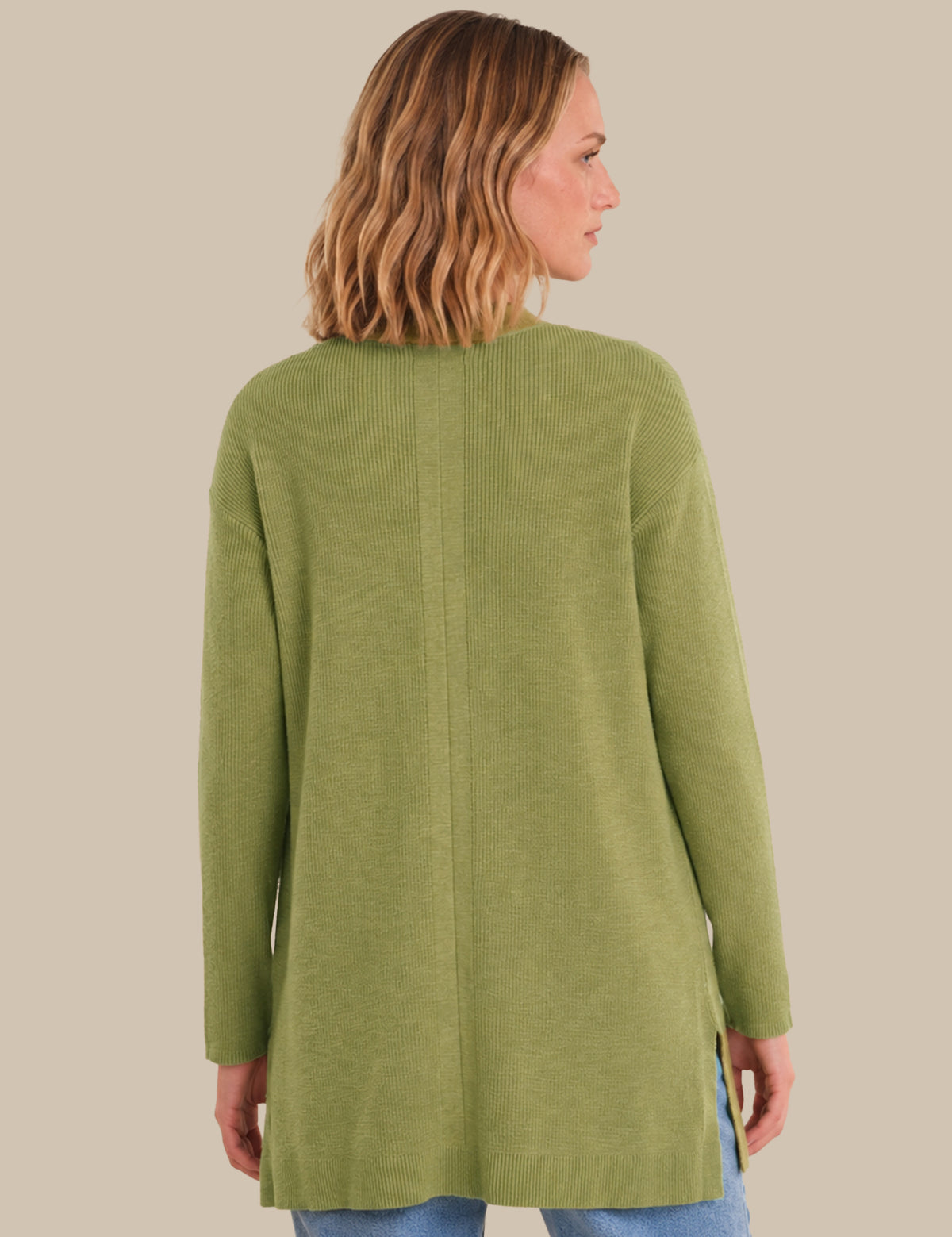 A woman shown from the back wearing a sage green knit long-sleeve tunic sweater, paired with blue jeans, against a solid beige background.