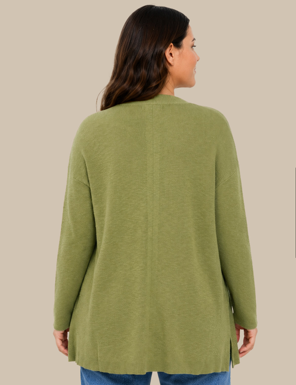 A woman viewed from the back wearing an olive green knit cardigan sweater with side slits, paired with blue jeans, against a solid beige background.