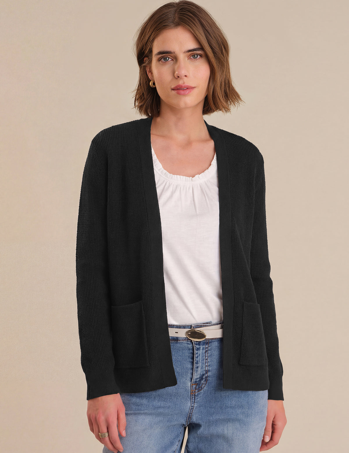 Woman wearing a black cardigan over a white shirt and blue jeans on a beige background