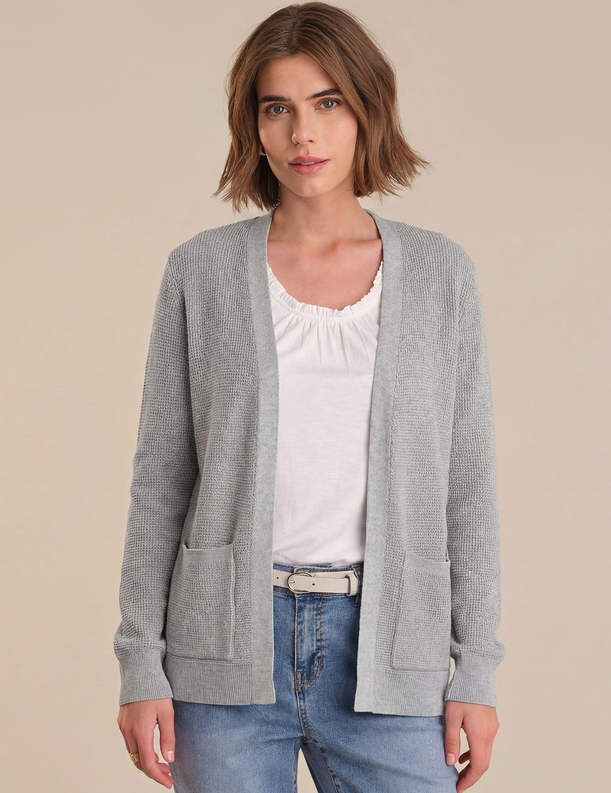 Woman wearing a gray cardigan over a white shirt and blue jeans on a beige background